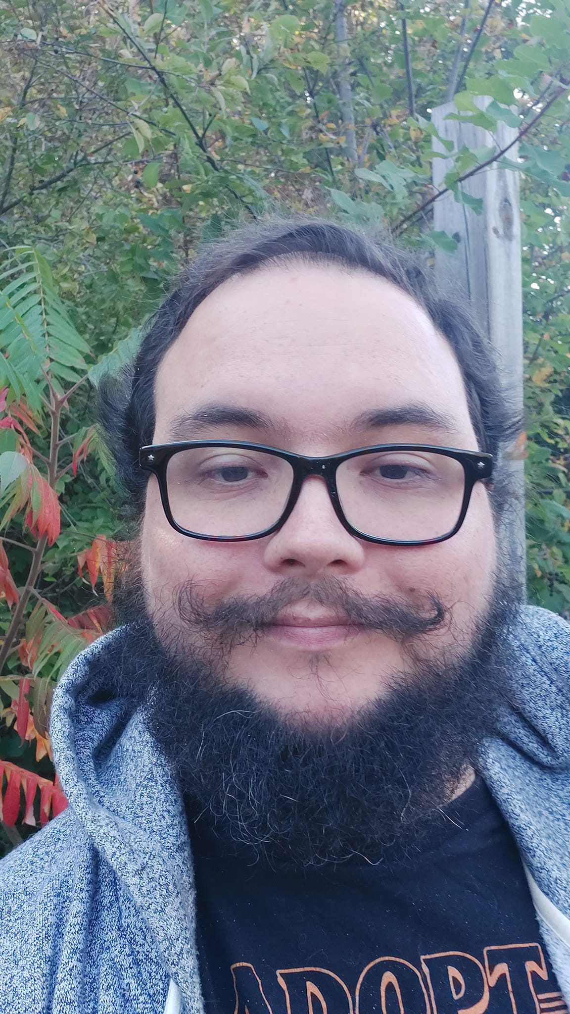 A man with short hair and bushy facial hair stands in front of a sumac bush which has begun to turn red in the autumn, his moustache curls on the ends